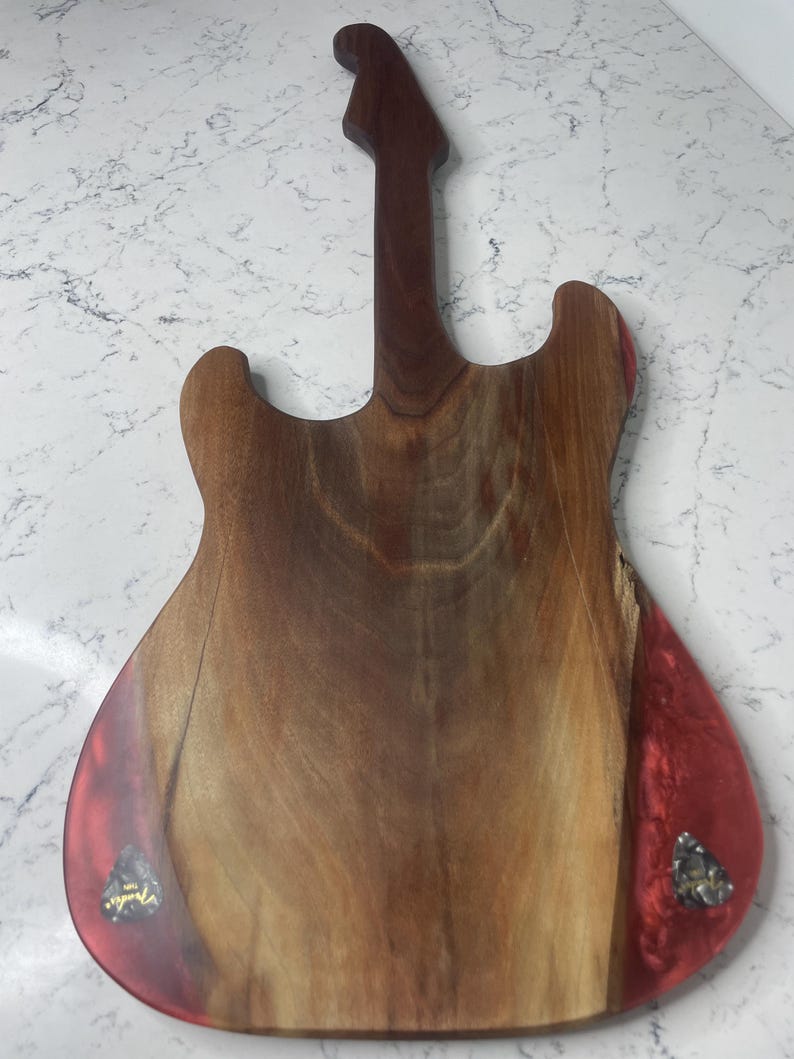 May include: A wooden guitar-shaped cutting board with a dark brown neck and body. The body has a reddish-brown hue with a red resin accent on the edges. Two guitar picks are embedded in the resin.