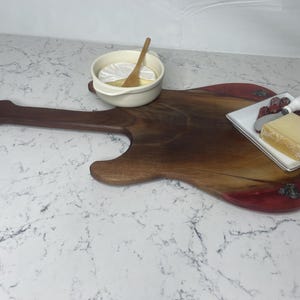 May include: A guitar-shaped wooden serving board with a dark brown finish and a red-toned edge. A small white bowl with a wooden spoon and a plate with cheese and fruit are on the board. The board is on a white marble surface.