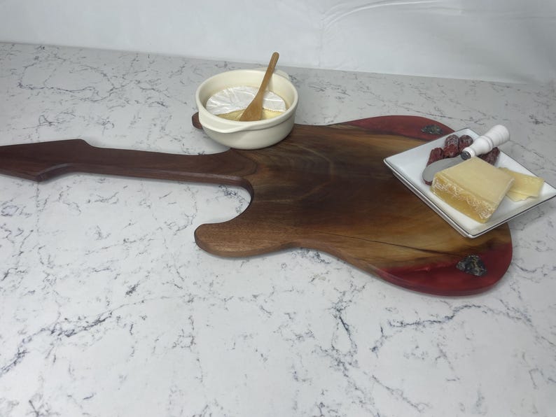 May include: A guitar-shaped wooden serving board with a dark brown finish and a red-toned edge. A small white bowl with a wooden spoon and a plate with cheese and fruit are on the board. The board is on a white marble surface.