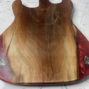 May include: A wooden guitar-shaped cutting board with a natural wood grain center and red resin edges. Two guitar picks are embedded in the resin. The board is designed for kitchen use and adds a unique touch to food preparation.