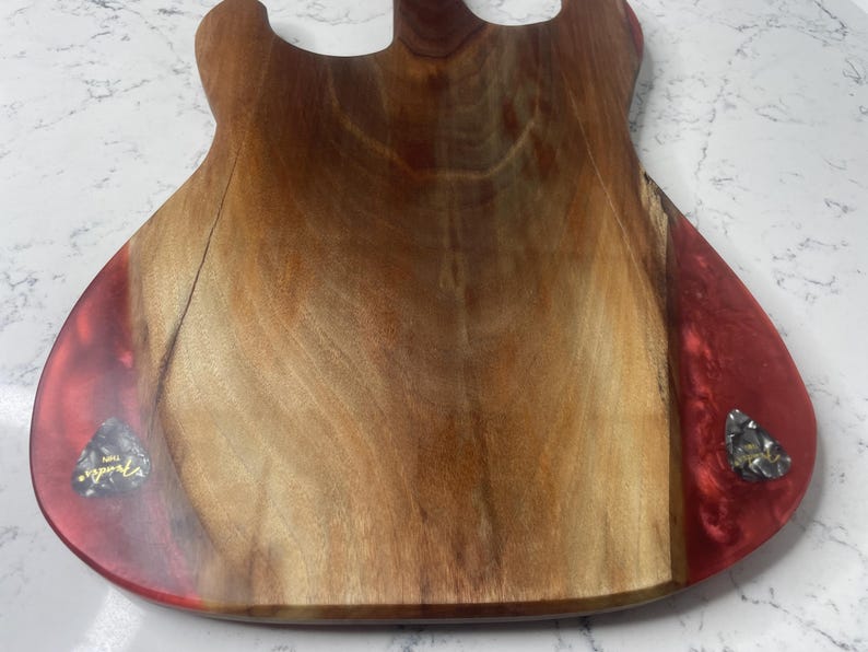 May include: A wooden guitar-shaped cutting board with a natural wood grain center and red resin edges. Two guitar picks are embedded in the resin. The board is designed for kitchen use and adds a unique touch to food preparation.