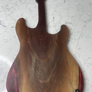May include: A wooden guitar-shaped cutting board with a dark brown neck and a lighter brown body. The body has a reddish-brown accent on the edges and is secured with black hardware. The wood grain is visible.