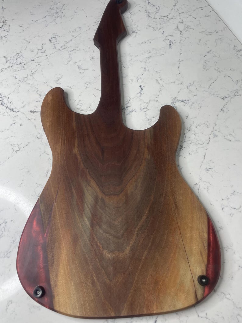 May include: A wooden guitar-shaped cutting board with a dark brown neck and a lighter brown body. The body has a reddish-brown accent on the edges and is secured with black hardware. The wood grain is visible.