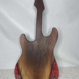 May include: A wooden guitar-shaped cutting board with a dark brown neck and body. The body features a natural wood grain pattern with red resin accents. The board is supported by a wooden stand.