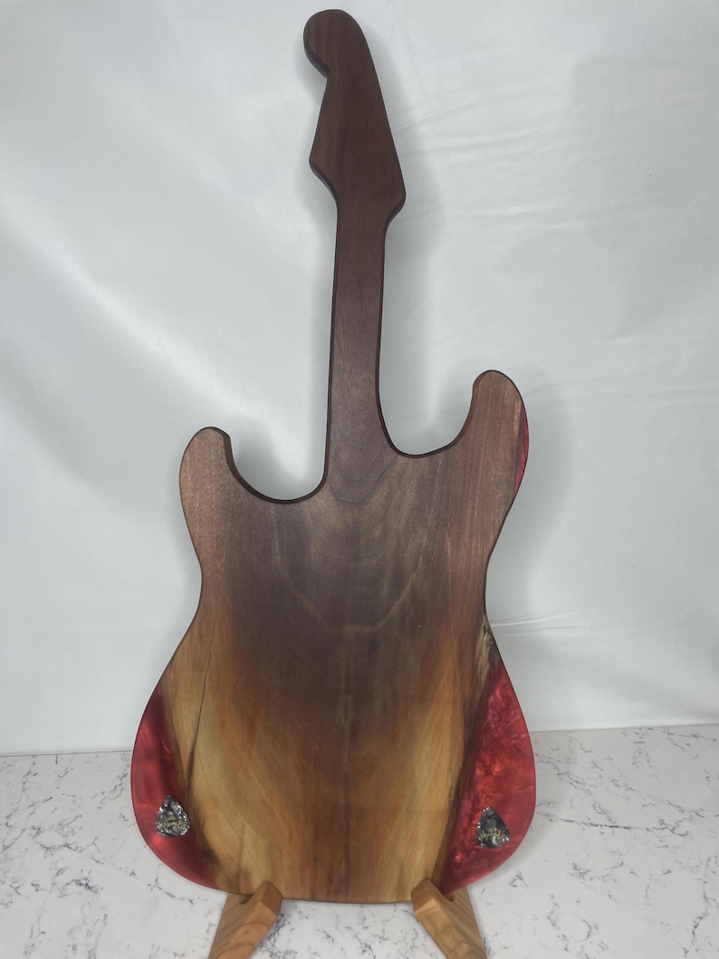 May include: A wooden guitar-shaped cutting board with a dark brown neck and body. The body features a natural wood grain pattern with red resin accents. The board is supported by a wooden stand.