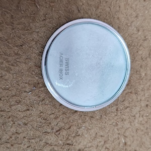 May include: A round, silver-colored watch back with a light blue face. The text "SWISS ACIER INOX" is printed on the face. The watch back is on a brown surface.
