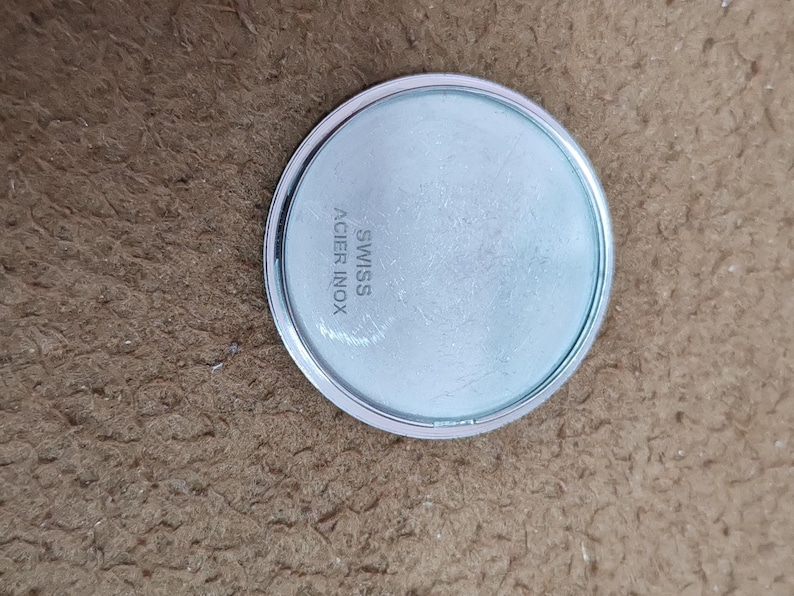 May include: A round, silver-colored watch back with a light blue face. The text "SWISS ACIER INOX" is printed on the face. The watch back is on a brown surface.