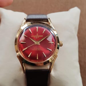 May include: A gold-tone Ulysse Nardin wristwatch featuring a vibrant red dial with a textured vertical striped pattern, gold-tone hour markers, and gold hands. The watch includes a dark brown leather strap and displays the text Ulysse Nardin, 17 Jewels, and Shock Proof on the face.
