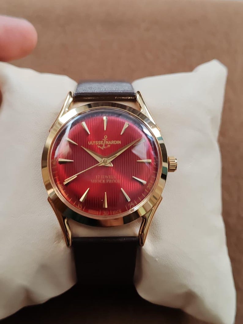 May include: A gold-tone Ulysse Nardin wristwatch featuring a vibrant red dial with a textured vertical striped pattern, gold-tone hour markers, and gold hands. The watch includes a dark brown leather strap and displays the text Ulysse Nardin, 17 Jewels, and Shock Proof on the face.