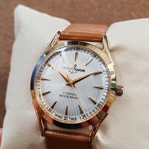 May include: A gold-tone wristwatch with a white, vertically textured dial, gold hands, and gold hour markers. The dial features the text ULYSSE NARDIN, 17 JEWELS, SHOCK PROOF, and SWISS MADE. The watch has a tan leather strap and a gold-tone crown.