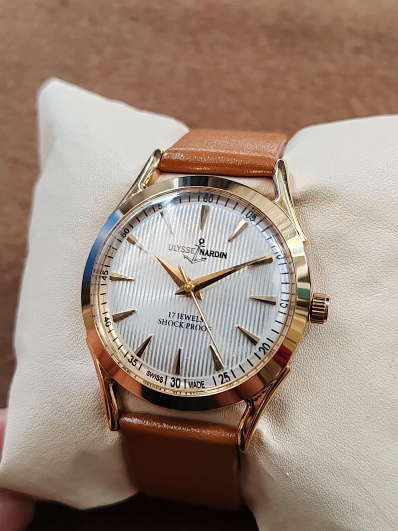 May include: A gold-tone wristwatch with a white, vertically textured dial, gold hands, and gold hour markers. The dial features the text ULYSSE NARDIN, 17 JEWELS, SHOCK PROOF, and SWISS MADE. The watch has a tan leather strap and a gold-tone crown.