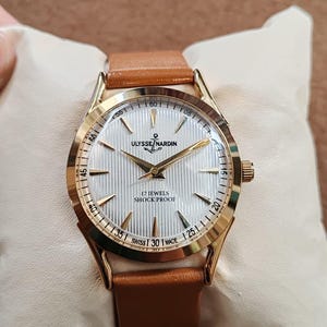 May include: A gold-tone wristwatch with a white, vertically textured dial, gold hands, and gold hour markers. The dial features the text ULYSSE NARDIN, an anchor logo, 17 JEWELS, SHOCK PROOF, and SWISS MADE. It has a tan leather strap.