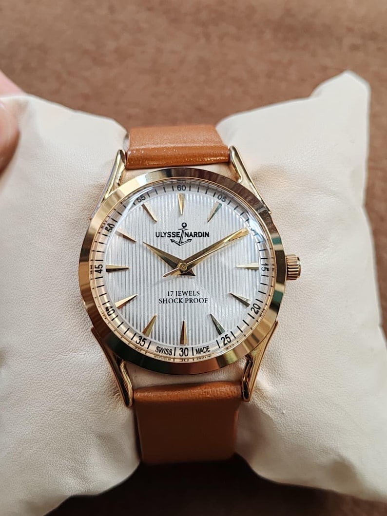 May include: A gold-tone wristwatch with a white, vertically textured dial, gold hands, and gold hour markers. The dial features the text ULYSSE NARDIN, an anchor logo, 17 JEWELS, SHOCK PROOF, and SWISS MADE. It has a tan leather strap.