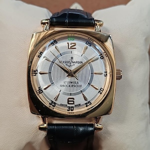 May include: A gold-tone square wristwatch with a black leather strap. The silver dial features a vertical striped pattern, gold-tone hands, and indices. Text on the dial reads Ulysse Nardin, 17 Jewels, Shock Proof, and Swiss Made, with numerals 12 and 6.