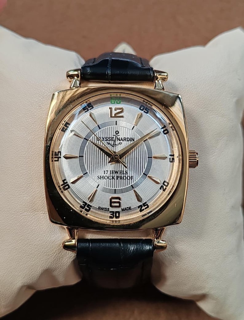 May include: A gold-tone square wristwatch with a black leather strap. The silver dial features a vertical striped pattern, gold-tone hands, and indices. Text on the dial reads Ulysse Nardin, 17 Jewels, Shock Proof, and Swiss Made, with numerals 12 and 6.