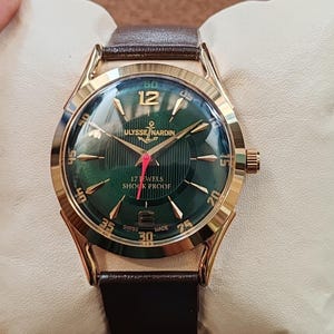 May include: A gold-tone wristwatch with a dark green sunburst dial, featuring gold-tone hour markers, a red second hand, and a brown leather strap. The dial displays the text ULYSSE NARDIN, 17 JEWELS, SHOCK PROOF, and SWISS MADE.