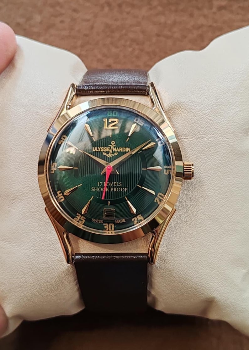 May include: A gold-tone wristwatch with a dark green sunburst dial, featuring gold-tone hour markers, a red second hand, and a brown leather strap. The dial displays the text ULYSSE NARDIN, 17 JEWELS, SHOCK PROOF, and SWISS MADE.