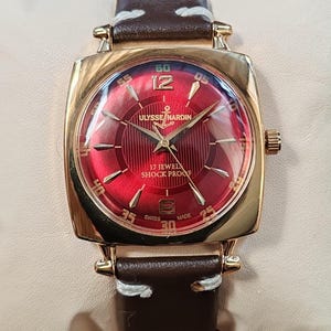 May include: A gold-tone wristwatch with a circular red dial featuring a textured pattern, gold-tone hands, and indices. The dial displays the text ULYSSE NARDIN, 17 JEWELS, SHOCK PROOF, and SWISS MADE. It has a brown leather strap with white stitching.