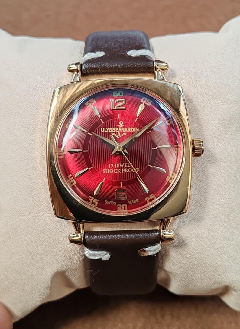 May include: A gold-tone wristwatch with a circular red dial featuring a textured pattern, gold-tone hands, and indices. The dial displays the text ULYSSE NARDIN, 17 JEWELS, SHOCK PROOF, and SWISS MADE. It has a brown leather strap with white stitching.