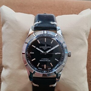 K&ouml;nnte beinhalten: Eine Armbanduhr mit schwarzem Lederarmband und silberfarbener geriffelter L&uuml;nette. Das schwarze Zifferblatt zeigt silberfarbene Zeiger und Indizes sowie den Schriftzug ULYSSE NARDIN, 17 JEWELS, SHOCK PROOF und SWISS MADE. Das Geh&auml;use ist aus Edelstahl.