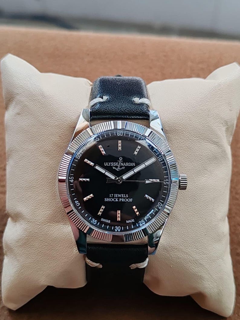 K&ouml;nnte beinhalten: Eine Armbanduhr mit schwarzem Lederarmband und silberfarbener geriffelter L&uuml;nette. Das schwarze Zifferblatt zeigt silberfarbene Zeiger und Indizes sowie den Schriftzug ULYSSE NARDIN, 17 JEWELS, SHOCK PROOF und SWISS MADE. Das Geh&auml;use ist aus Edelstahl.