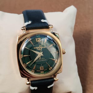 K&ouml;nnte beinhalten: Eine goldfarbene quadratische Armbanduhr mit dunkelgr&uuml;nem Zifferblatt und der Aufschrift Ulysse Nardin, 17 Jewels, Shock Proof und Swiss Made. Die Uhr hat goldfarbene Zeiger und Indizes sowie ein schwarzes Lederarmband mit wei&szlig;en N&auml;hten.