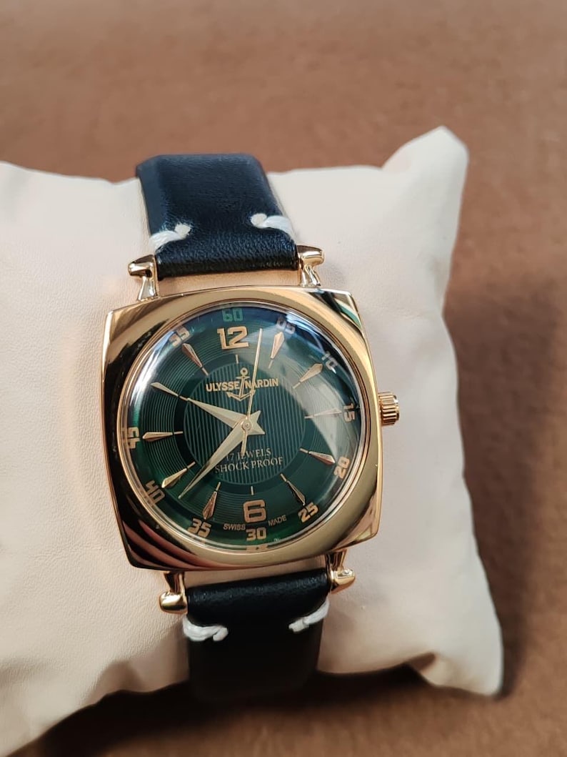 K&ouml;nnte beinhalten: Eine goldfarbene quadratische Armbanduhr mit dunkelgr&uuml;nem Zifferblatt und der Aufschrift Ulysse Nardin, 17 Jewels, Shock Proof und Swiss Made. Die Uhr hat goldfarbene Zeiger und Indizes sowie ein schwarzes Lederarmband mit wei&szlig;en N&auml;hten.
