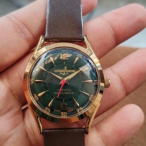 May include: A gold-tone wristwatch with a dark green dial featuring vertical pinstripes, gold-tone hour markers, and the text Ulysse Nardin, 17 Jewels, Shock Proof, and Swiss Made. The watch has a brown leather strap and a red second hand.
