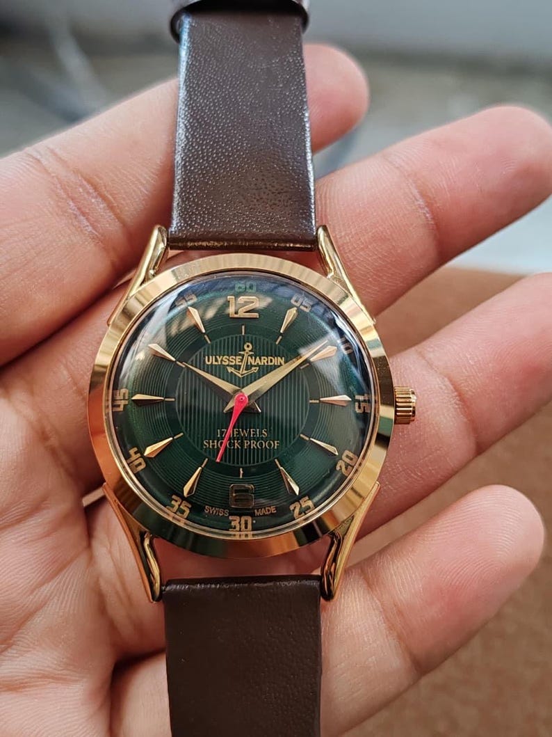 May include: A gold-tone wristwatch with a dark green dial featuring vertical pinstripes, gold-tone hour markers, and the text Ulysse Nardin, 17 Jewels, Shock Proof, and Swiss Made. The watch has a brown leather strap and a red second hand.