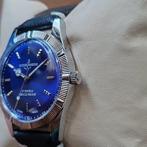 May include: A silver-tone wristwatch with a deep blue sunburst dial, featuring the text ULYSSE NARDIN, 17 JEWELS, SHOCK PROOF, and SWISS MADE. The watch has a fluted bezel, silver hour markers, and a black leather strap with white stitching.