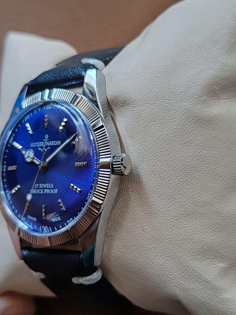 May include: A silver-tone wristwatch with a deep blue sunburst dial, featuring the text ULYSSE NARDIN, 17 JEWELS, SHOCK PROOF, and SWISS MADE. The watch has a fluted bezel, silver hour markers, and a black leather strap with white stitching.