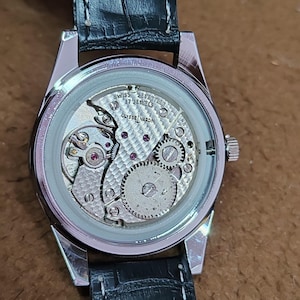 May include: A silver-toned wristwatch with a black leather band. The watch's back is transparent, revealing the intricate internal mechanism, including gears, jewels, and the text "SWISS 17 JEWELS ULYSSE NARDIN".