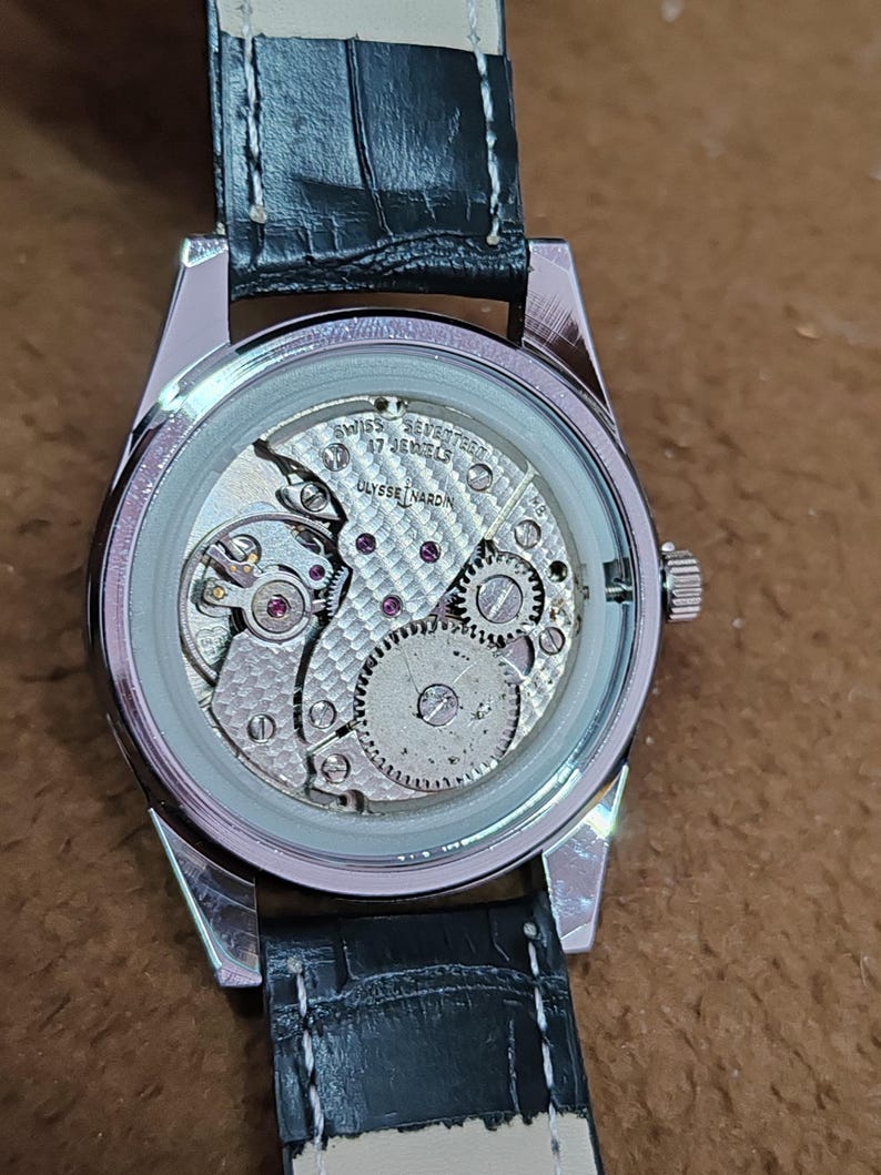 May include: A silver-toned wristwatch with a black leather band. The watch's back is transparent, revealing the intricate internal mechanism, including gears, jewels, and the text "SWISS 17 JEWELS ULYSSE NARDIN".