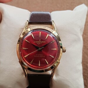 May include: A gold-tone wristwatch with a deep red dial featuring vertical pinstripes, gold-tone hands, and hour markers. The dial displays the text ULYSSE NARDIN with an anchor logo, 17 JEWELS, and SHOCK PROOF. It has a dark brown leather strap.