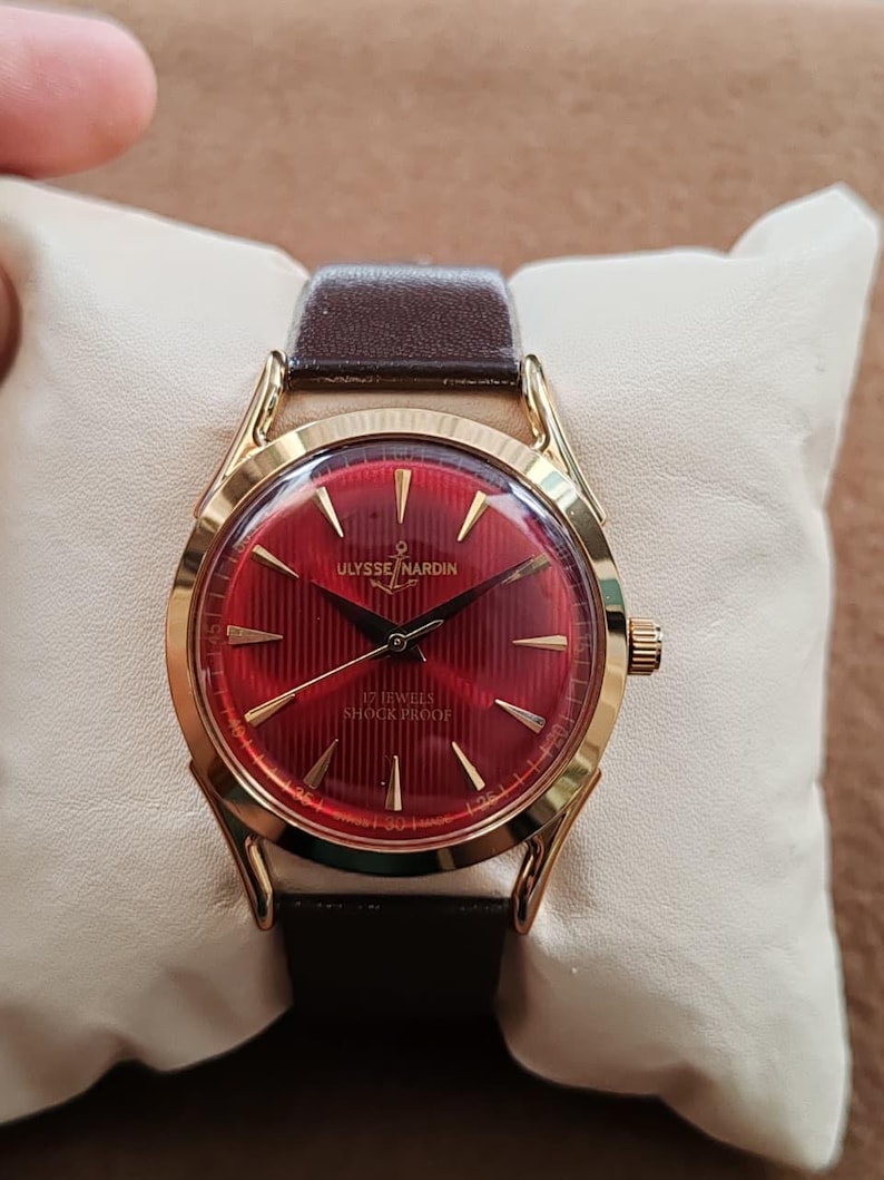 May include: A gold-tone wristwatch with a deep red dial featuring vertical pinstripes, gold-tone hands, and hour markers. The dial displays the text ULYSSE NARDIN with an anchor logo, 17 JEWELS, and SHOCK PROOF. It has a dark brown leather strap.