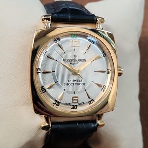 May include: A gold-tone wristwatch with a square case and a black leather strap. The silver dial features gold-tone hour markers, the text ULYSSE NARDIN with an anchor logo, 17 JEWELS, SHOCK PROOF, and SWISS MADE. The watch has gold-tone hands.