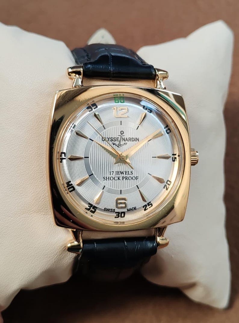 May include: A gold-tone wristwatch with a square case and a black leather strap. The silver dial features gold-tone hour markers, the text ULYSSE NARDIN with an anchor logo, 17 JEWELS, SHOCK PROOF, and SWISS MADE. The watch has gold-tone hands.