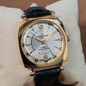 May include: A gold-tone wristwatch with a square case and a black leather strap. The silver dial features a textured pattern, gold-tone hands, Arabic numerals at 12 and 6, and the text Ulysse Nardin, 17 Jewels, Shock Proof, and Swiss Made.