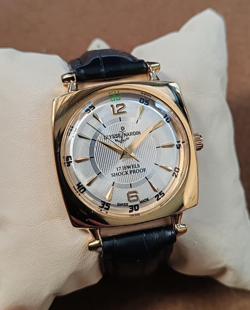 May include: A gold-tone wristwatch with a square case and a black leather strap. The silver dial features a textured pattern, gold-tone hands, Arabic numerals at 12 and 6, and the text Ulysse Nardin, 17 Jewels, Shock Proof, and Swiss Made.