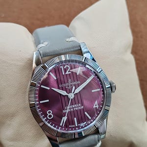 May include: A wristwatch with a round silver-tone case and a fluted bezel. The dial is a deep magenta color with vertical stripes, white Arabic numerals at 12, 4, and 8, and the text Ulysse Nardin, 17 Jewels, Shock Proof, and Swiss Made.