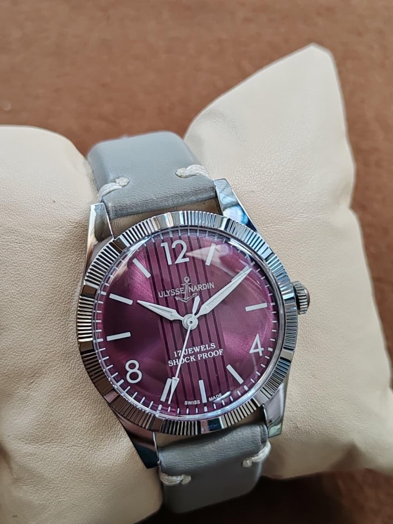 May include: A wristwatch with a round silver-tone case and a fluted bezel. The dial is a deep magenta color with vertical stripes, white Arabic numerals at 12, 4, and 8, and the text Ulysse Nardin, 17 Jewels, Shock Proof, and Swiss Made.
