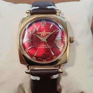 May include: A gold-tone wristwatch with a square case and a deep red sunburst dial. The dial features gold-tone indices, the text ULYSSE NARDIN, 17 JEWELS, SHOCK PROOF, and SWISS MADE. It has a dark brown leather strap with white stitching.