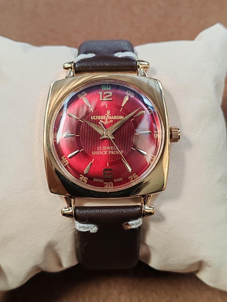 May include: A gold-tone wristwatch with a square case and a deep red sunburst dial. The dial features gold-tone indices, the text ULYSSE NARDIN, 17 JEWELS, SHOCK PROOF, and SWISS MADE. It has a dark brown leather strap with white stitching.