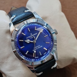 May include: A silver-tone wristwatch with a deep blue dial, featuring the text ULYSSE NARDIN, 17 JEWELS, SHOCK PROOF, and SWISS MADE. The watch has a fluted bezel, silver hands, and a black leather strap with white stitching.