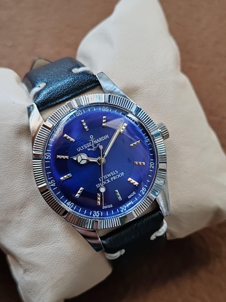 May include: A silver-tone wristwatch with a deep blue dial, featuring the text ULYSSE NARDIN, 17 JEWELS, SHOCK PROOF, and SWISS MADE. The watch has a fluted bezel, silver hands, and a black leather strap with white stitching.