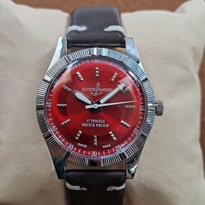 May include: A wristwatch with a dark brown leather strap and a silver-toned fluted bezel. The watch features a vibrant red dial with silver hour markers, white hands, and the text Ulysse Nardin, 17 Jewels, Shock Proof, and Swiss Made.
