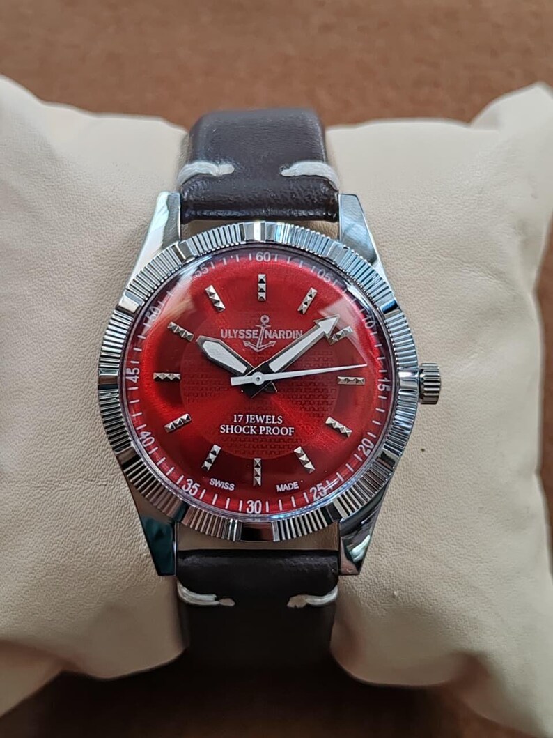 May include: A wristwatch with a dark brown leather strap and a silver-toned fluted bezel. The watch features a vibrant red dial with silver hour markers, white hands, and the text Ulysse Nardin, 17 Jewels, Shock Proof, and Swiss Made.