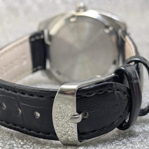 May include: A wristwatch featuring a black leather strap with a textured crocodile pattern and a silver-toned metal buckle. The watch case is circular with a polished silver-toned metal back, and the strap interior is lined with a light beige material.