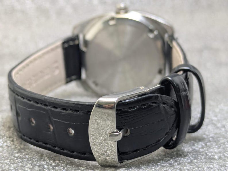 May include: A wristwatch featuring a black leather strap with a textured crocodile pattern and a silver-toned metal buckle. The watch case is circular with a polished silver-toned metal back, and the strap interior is lined with a light beige material.