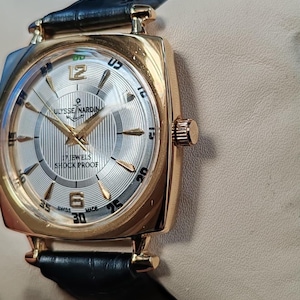 May include: A gold-tone Ulysse Nardin wristwatch with a square case and a black leather strap. The silver dial features a striped pattern, gold-tone hands, gold-tone hour markers, and the text Ulysse Nardin, 17 Jewels, Shock Proof, and Swiss Made.
